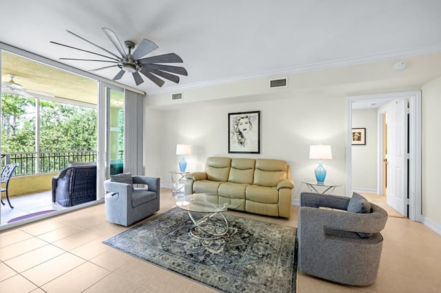 Bright condo living room with tile floors, large ceiling fan, gray armchairs and sliding doors to a screened lanai