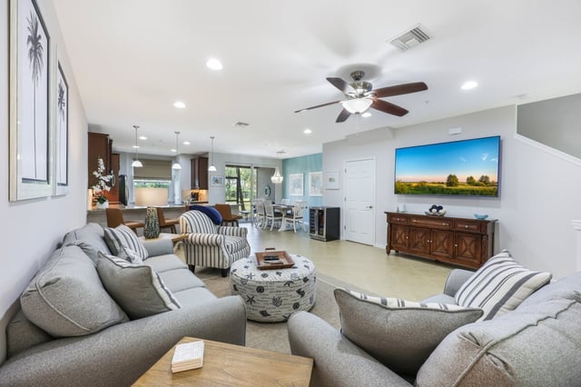 Open-concept living room with gray sectional sofas, ceiling fan, wall-mounted TV and kitchen beyond