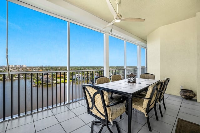 Screened high-rise balcony with a tiled dining table, six cushioned chairs and ceiling fan overlooking a waterway and blue sky