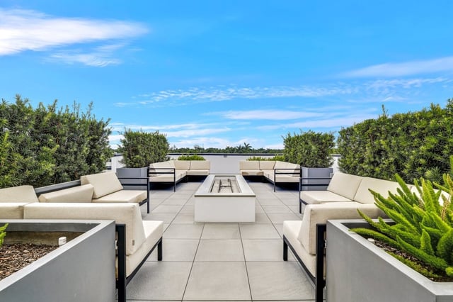 Rooftop terrace with white cushioned lounge seating around a linear fire pit, framed by hedges under blue sky