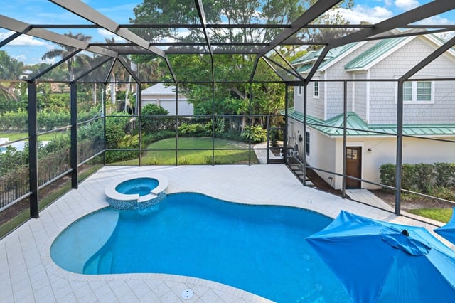 Screened lanai with a freeform pool, raised spa and blue umbrella beside a shingled home with green metal roof