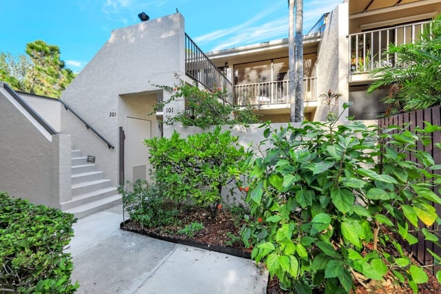 Entrance walkway to a two-story stucco condo building with exterior stairs, unit numbers 101 and 201, and lush green shrubs