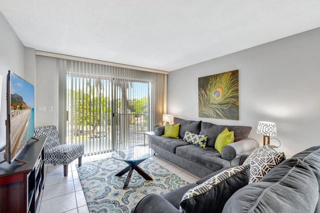 Bright condo living room with gray sofas, peacock feather artwork and sliding glass doors to a sunny patio