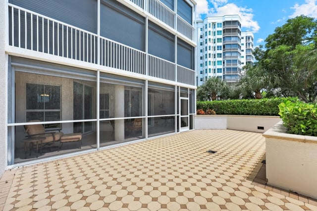 Spacious tiled patio beside a screened lanai with a high-rise condo tower and hedges in the background