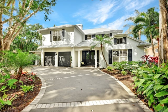 White two-story home with black shutters, a two-car garage and curving driveway framed by palms and tropical plants