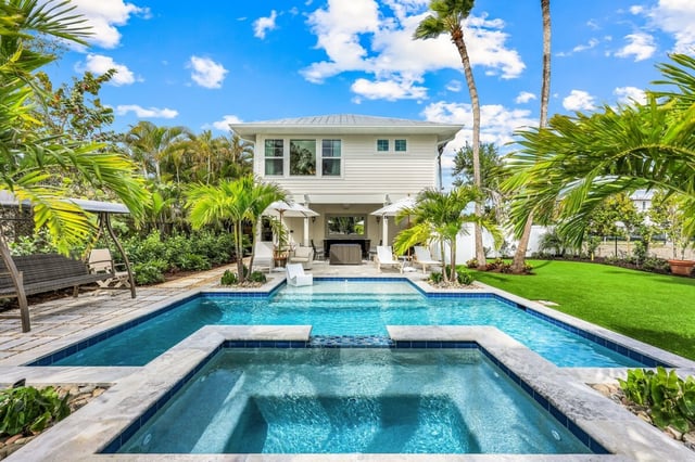 Backyard pool and spa with spillover edge framed by palms and a white two-story coastal home under blue sky