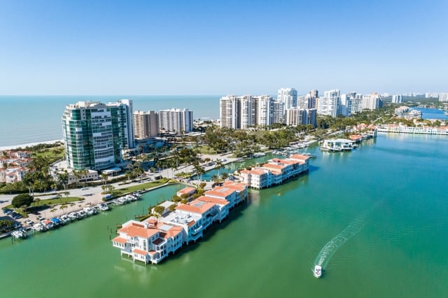 Aerial view of waterfront condo towers and red-roofed villas along a turquoise Naples bay with a boat leaving a wake
