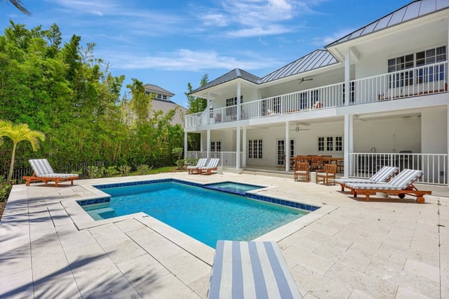 White two-story coastal home with metal roof and balcony overlooking a pool with striped loungers on travertine deck