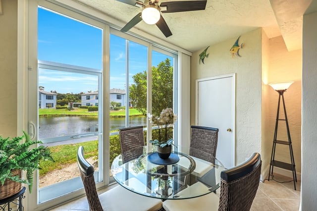 Sunlit dining nook with glass table, wicker chairs and sliding doors overlooking a lake and condos
```