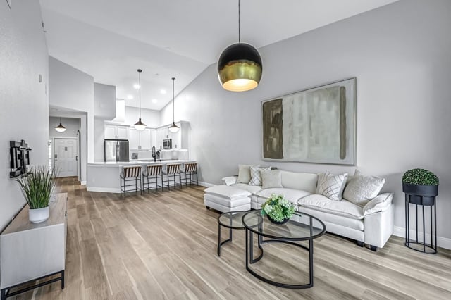 Bright open-plan living room with white sectional sofa, glass coffee tables, pendant lights and kitchen bar stools beyond