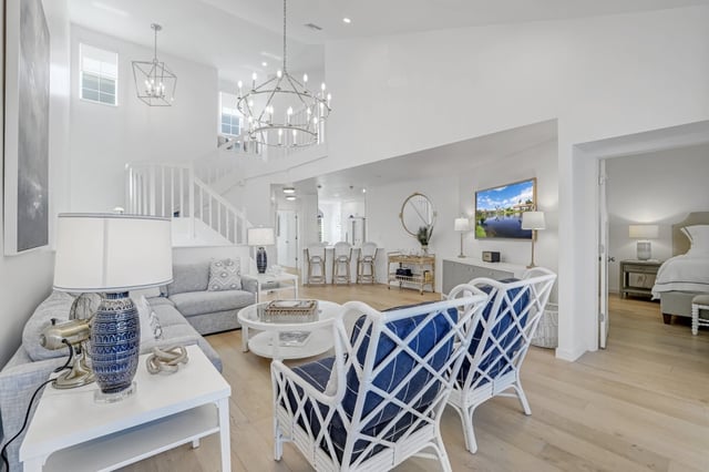 Bright white coastal living room with blue accent chairs, chandelier, staircase and open view to a bedroom