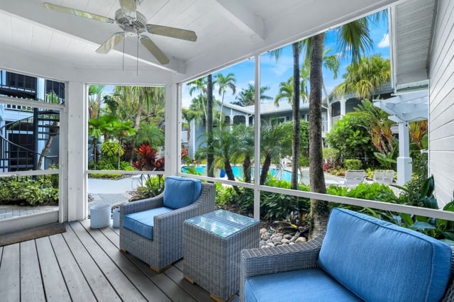 Screened porch with blue cushioned wicker chairs and ceiling fan overlooking a palm-lined pool courtyard