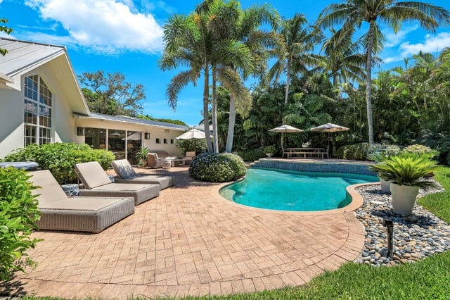 Curved backyard pool with wicker loungers, brick patio, patio umbrellas and tall palms under blue sky