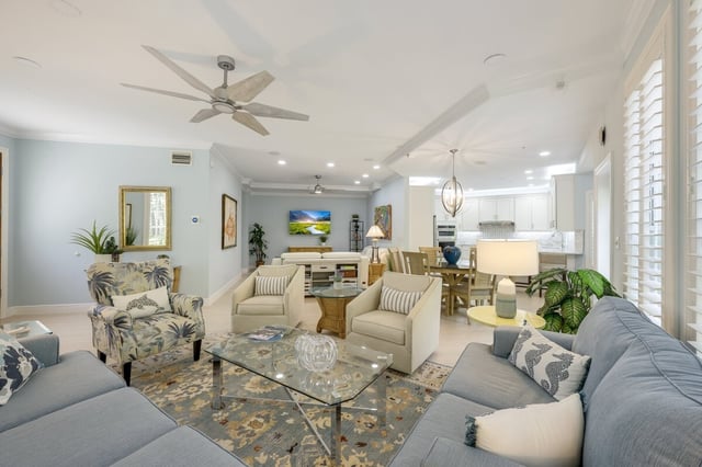 Open-plan coastal living room with gray sofas, armchairs, glass coffee table and ceiling fan leading to a dining area and kitchen