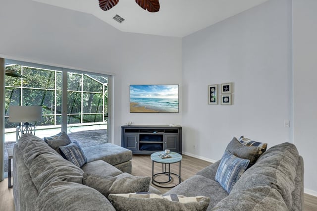Bright living room with gray sectional sofas, wall-mounted TV on a media console, and sliding doors to a screened lanai