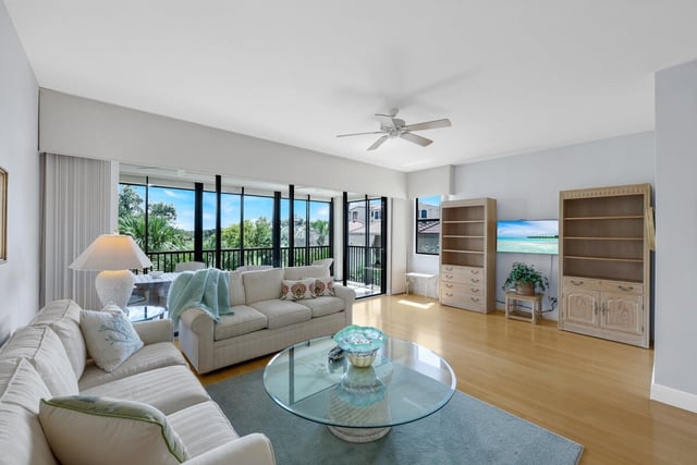 Bright coastal condo living room with cream sofas, glass coffee table, ceiling fan and windows overlooking greenery