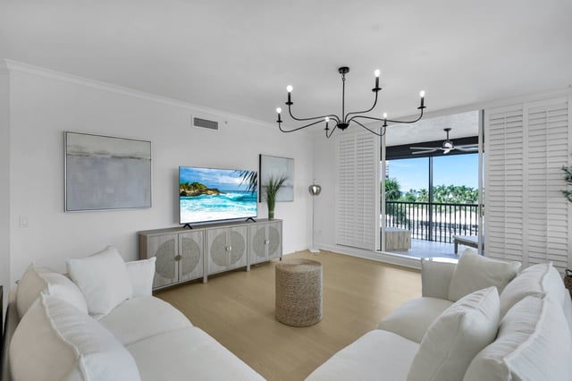 Bright white living room with sectional sofas, black chandelier, wall-mounted TV and balcony doors framing palm views