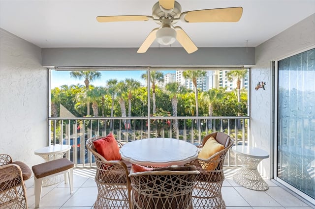 Screened condo balcony with wicker dining set, ceiling fan and palm treetop views over a pool