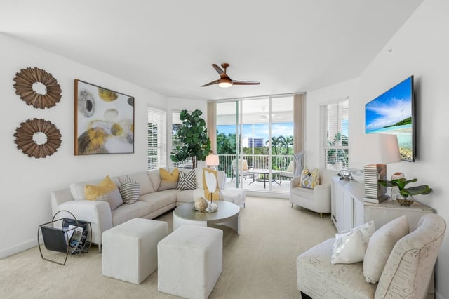 Bright coastal condo living room with cream sectional, ceiling fan, wall art, and balcony overlooking palm trees