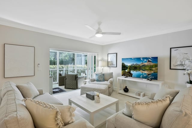 Bright coastal living room with white sofas, ceiling fan, wall-mounted TV and sliding doors to a green lanai