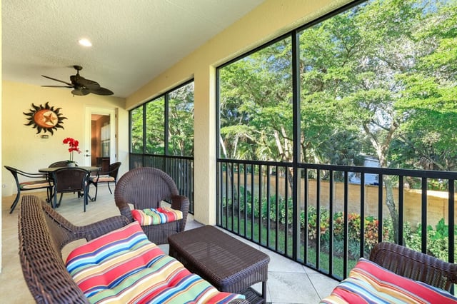 Screened lanai with striped wicker seating, ceiling fan and dining table overlooking lush green trees and garden