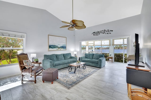 Bright vaulted living room with teal sofas, palm-leaf ceiling fan and sliding doors opening to a water view