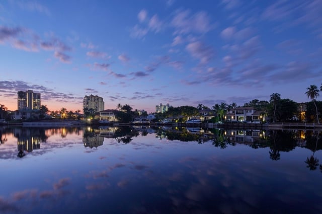 Twilight sky mirrored in glassy water beside waterfront homes, palm trees and high-rise towers with lit windows