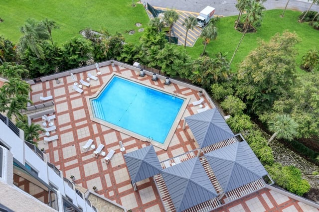 Aerial view of a blue rectangular pool with lounge chairs, patterned deck, gray cabanas and palm-filled lawns