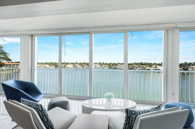 Bright condo living room with floor-to-ceiling windows overlooking a waterway, white round table and blue armchairs