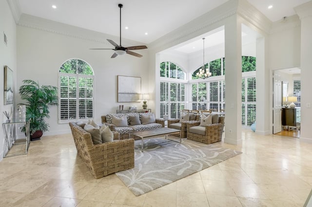 Bright high-ceilinged living room with wicker armchairs, arched shuttered windows and travertine tile floors