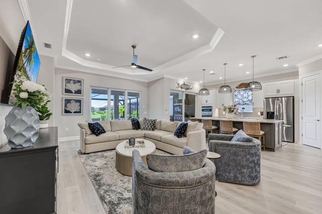 Open-concept living room with beige sectional, swivel armchairs, tray ceiling and adjoining kitchen with pendant lights