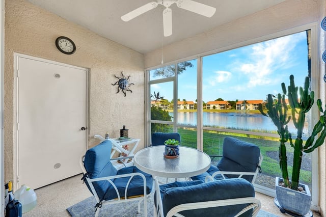 Screened lanai with blue cushioned patio chairs and glass table overlooking a sunlit lake and tile-roofed condos