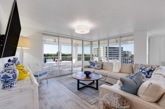 Bright condo living room with beige sectional sofas, round wood coffee table and floor-to-ceiling windows overlooking treetops
