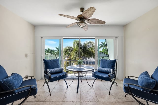 Sunlit sitting room with blue cushioned chairs, glass table and palm-leaf ceiling fan facing a balcony with palm views