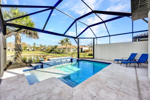 Screened lanai pool with water features, blue lounge chairs and travertine deck overlooking a lake and palm trees