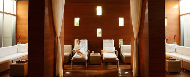 A woman in a white robe relaxes on a lounge chair in the warm wood-paneled relaxation room at The Spa at Naples Grande