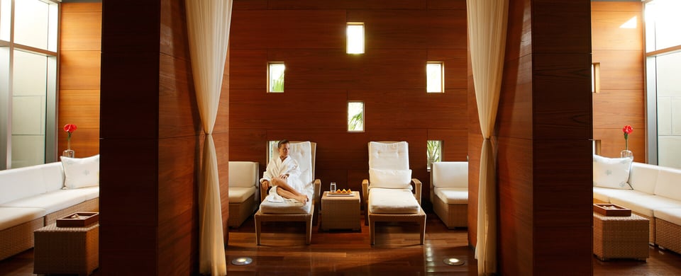 A woman in a white robe relaxes on a lounge chair in the warm wood-paneled relaxation room at The Spa at Naples Grande