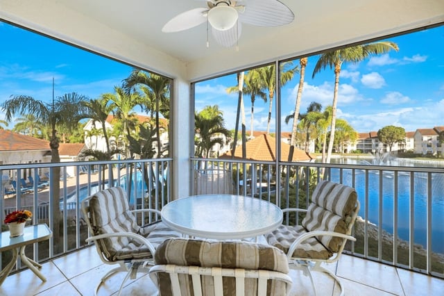 Screened lanai with a glass patio table and cushioned chairs overlooking a pool, palm trees and a lake fountain
