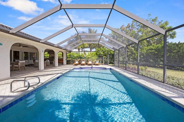 Screened lanai enclosing a long blue swimming pool beside a covered patio with lounge chairs and palm trees