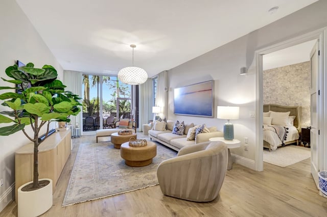 Bright coastal living room with neutral sofa, round leather ottomans, fiddle-leaf fig, and palm views through glass doors