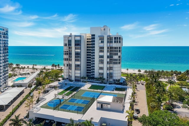 Aerial view of a white beachfront high-rise condo with tennis courts, pool and palms beside the turquoise Gulf
