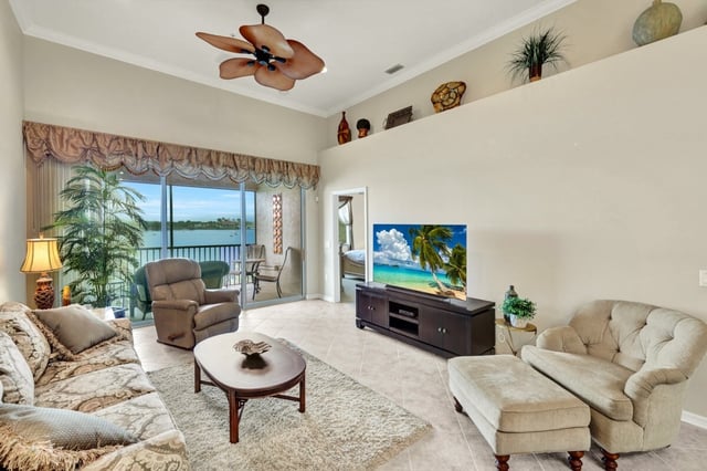 Bright condo living room with palm-leaf ceiling fan, flat-screen TV, and sliding doors opening to a water view balcony