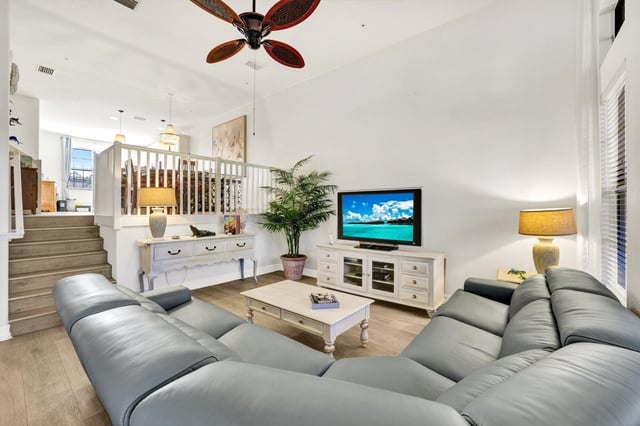 Bright two-story living room with gray sectional sofa, palm-leaf ceiling fan, TV console and white coffee table