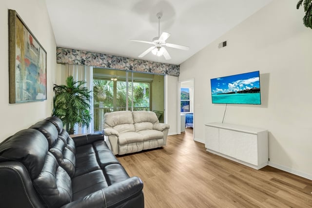 Bright living room with black leather sofa, beige loveseat, ceiling fan, wall-mounted TV and sliding doors to a lanai