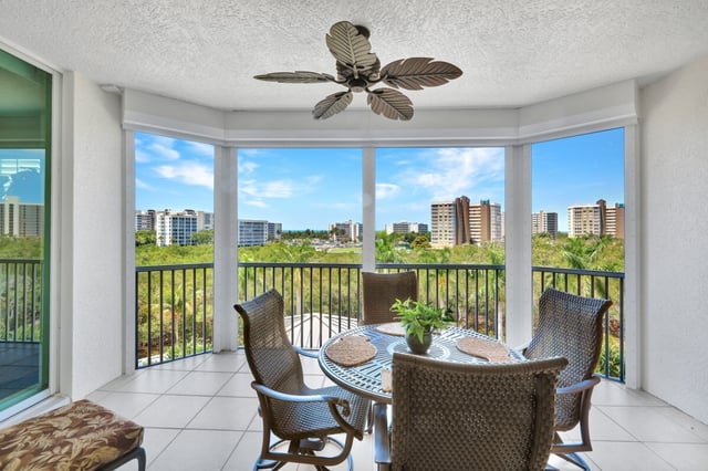 Screened condo balcony with wicker dining set, palm-leaf ceiling fan and views of high-rise towers and treetops