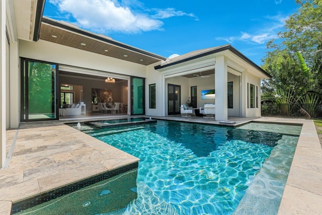 Turquoise backyard pool with spa and water features beside a modern home with open sliding glass walls and covered lanai