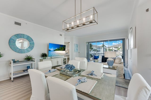 Bright coastal condo dining area with white slipcovered chairs, coral centerpiece, and living room opening to a palm-lined lanai