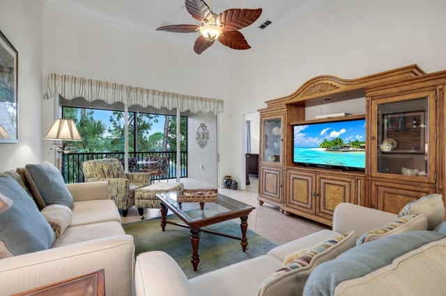 Bright condo living room with palm-leaf ceiling fan, wooden entertainment center and sofas facing a screened lanai