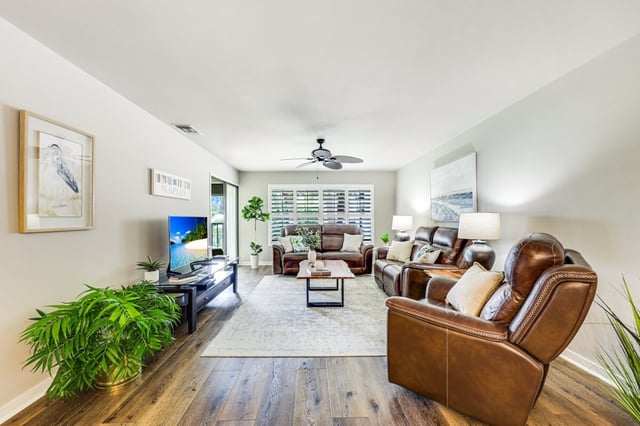 Bright living room with brown leather sofas, a ceiling fan, hardwood floors and potted palms near plantation shutters