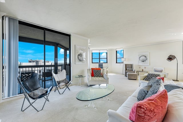 Bright white condo living room with butterfly chairs, glass coffee table and balcony doors opening to blue sky